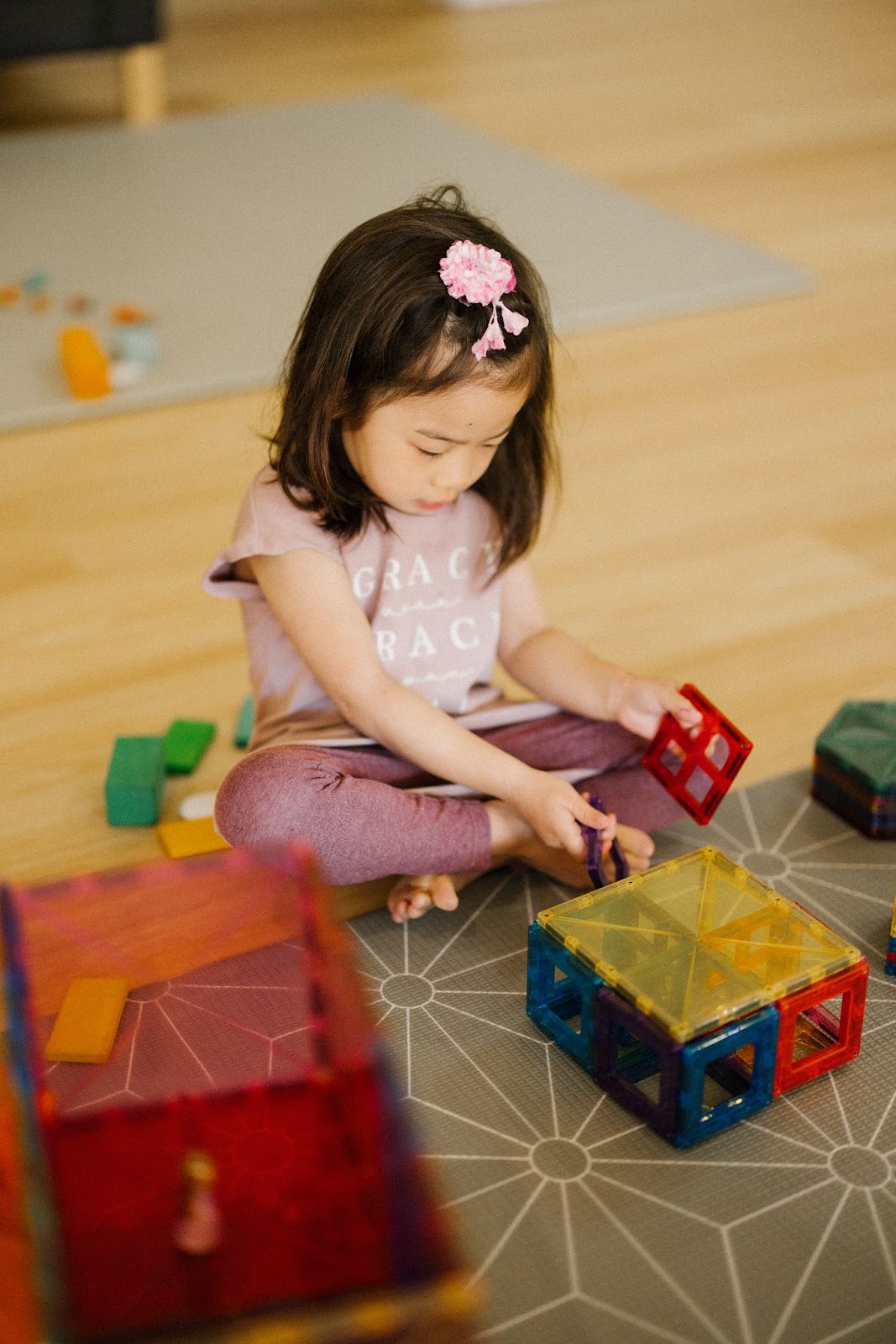 A toddler is focused on assembling magnetic tiles, using large and small square tiles to create cuboids in a colorful structure. The child carefully connects the pieces, developing hand-eye coordination and fine motor skills while exploring shapes and engaging in imaginative, hands-on play.
