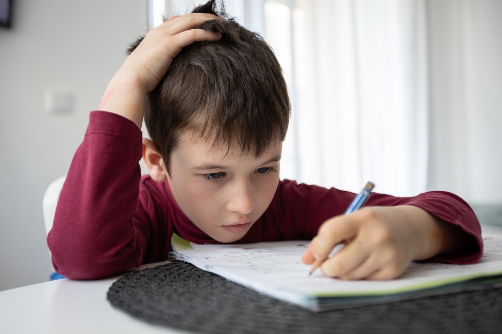 A young child scratches their head while doing homework at their kitchen table.