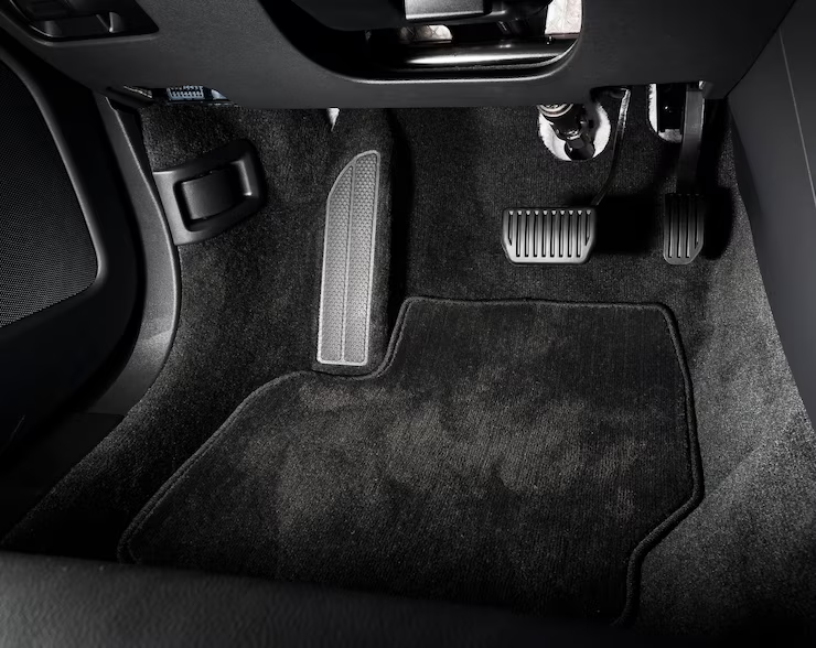 A black carpeted car floor mat installed in the driver's footwell, positioned under the accelerator, brake, and clutch pedals