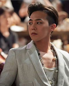 This contains an image of G-Dragon wearing a suit and necklace looking off into the distance with other people behind him