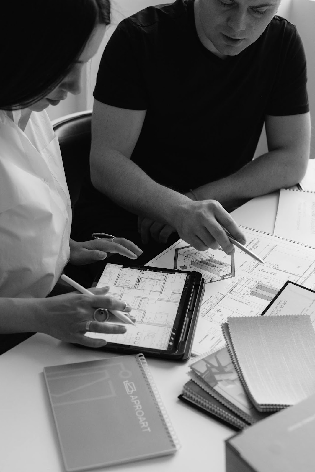 Two architects review architectural designs at a table. One uses a tablet for BIM 3D Design, while the other points at paper plans. Notebooks and folders are scattered on the table in this black and white scene.