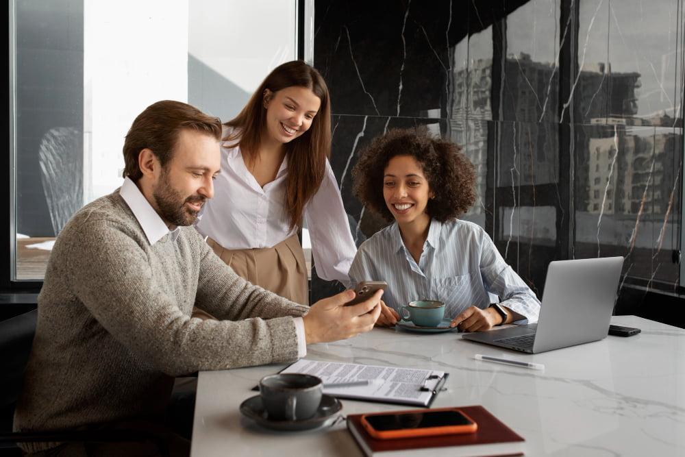 A team collaborating in a workspace, gathered around a mobile device and a laptop.