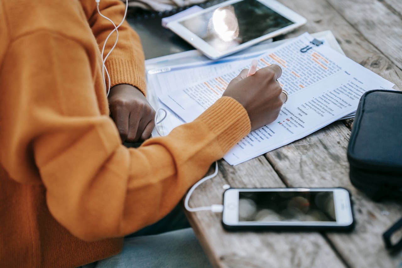 A person in a mustard sweater is studying outdoor materials with earphones, surrounded by a tablet, smartphone, and notes, focusing on learning strategies possibly for creating online courses.