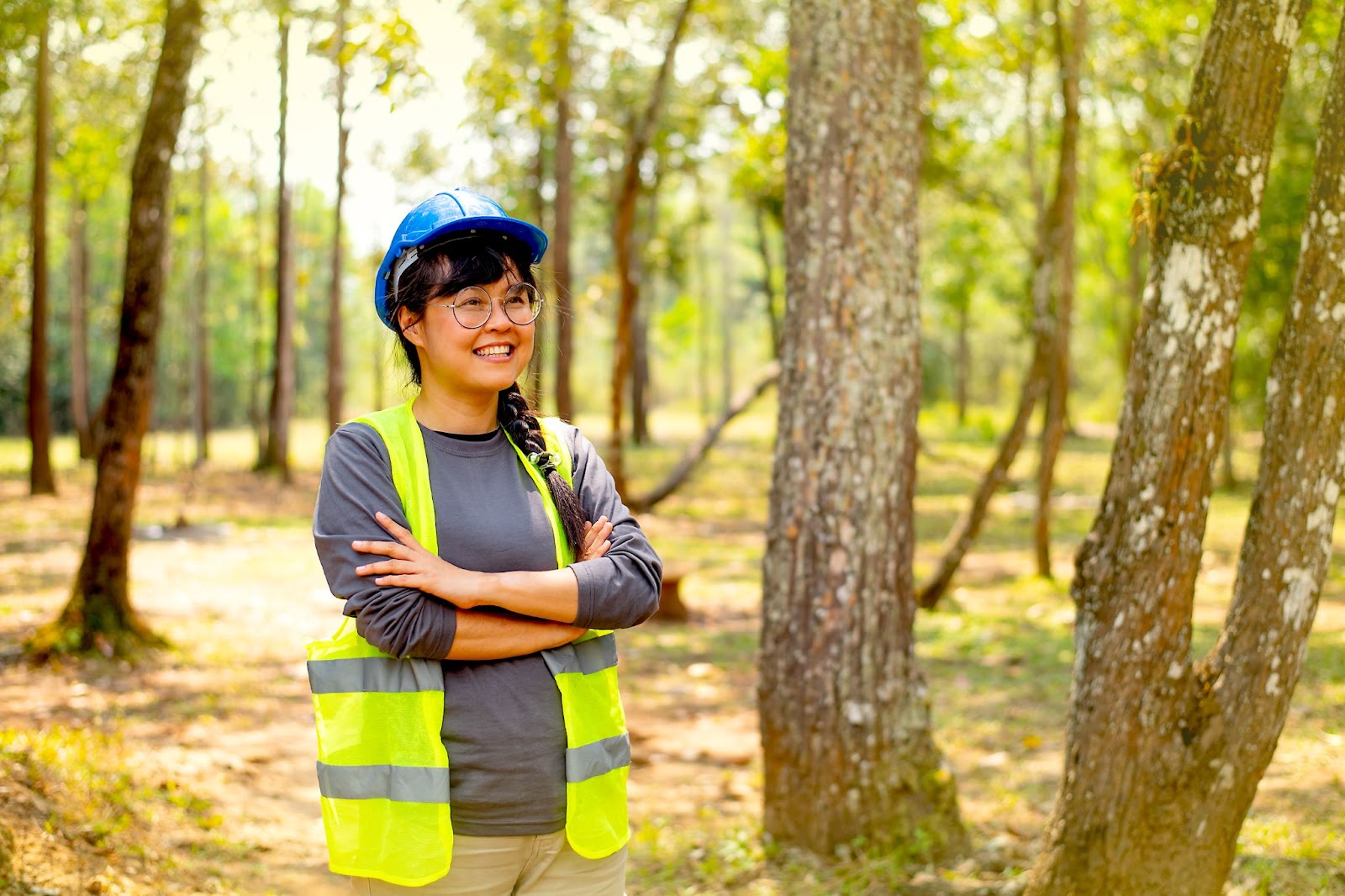 Woman in a hard hat and vest next to trees.