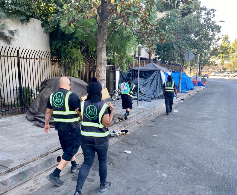 Four outreach workers from Urban Alchemy are walking down the street wearing all black and vests that identify them as part of Urban Alchemy, the nonprofit hired by Mitch O'Farrell's district to assist LAHSA with outreach. They are handing out food and water to unhoused people. 