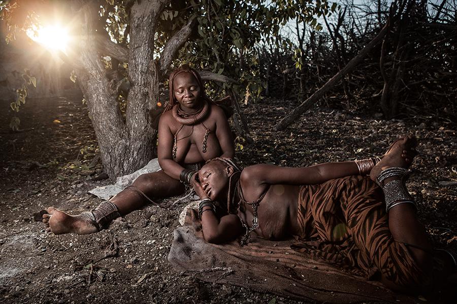 A picture of two Himba Women relaxing under a tree. One Himba woman applying otjize paste to the other woman's hair. Photographed by Ben McRae.