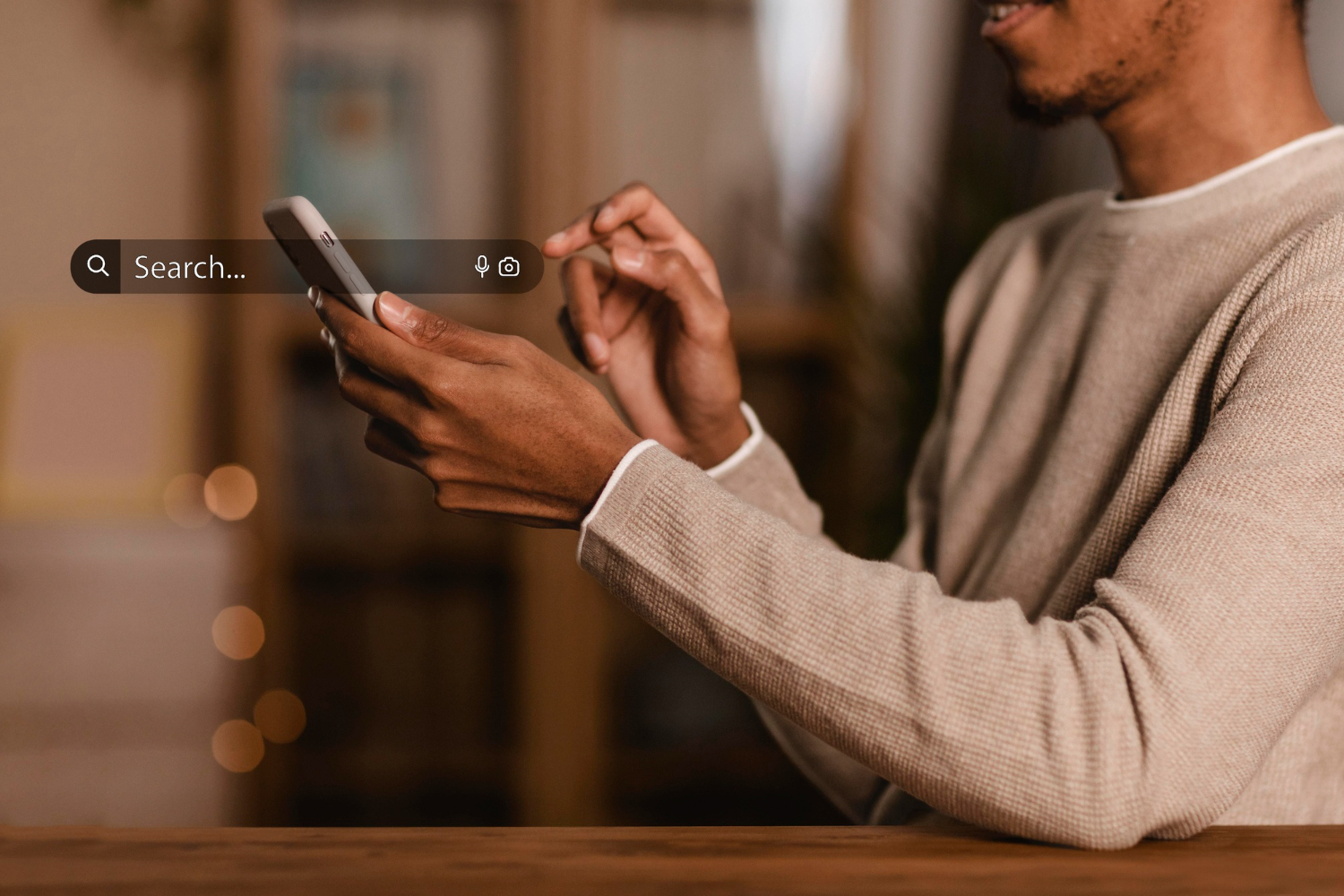 A imagem mostra um homem segurando um smartphone, com foco na interação digital, essencial para entender "como vender pela internet". Ele está em um ambiente acolhedor, com uma mesa de madeira ao fundo. O smartphone exibe uma barra de pesquisa, sugerindo que ele está buscando informações ou produtos online. A mão do homem está posicionada de forma a tocar a tela, indicando uma ação de pesquisa ou navegação. O ambiente é iluminado suavemente, criando uma atmosfera confortável e propícia para atividades digitais. Essa cena reflete a importância da tecnologia e da pesquisa na estratégia de vendas online.