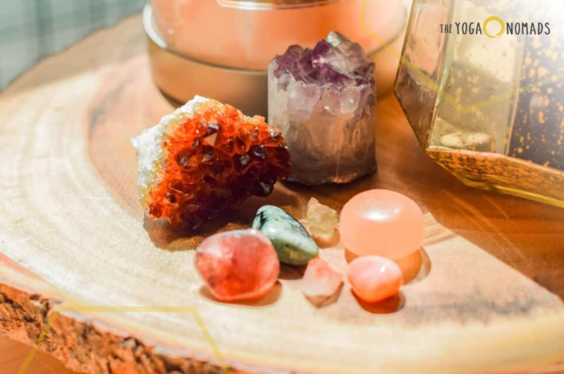 The image showcases a beautiful collection of crystals and stones arranged on a wooden surface. Prominently featured is a large amethyst geode, accompanied by a vibrant red-orange crystal, likely citrine. Surrounding these are smaller stones, including rose quartz and polished green and blue stones, possibly aventurine or turquoise. In the background, there’s a pinkish pot with a lid and a golden-colored decorative object with glass panels.