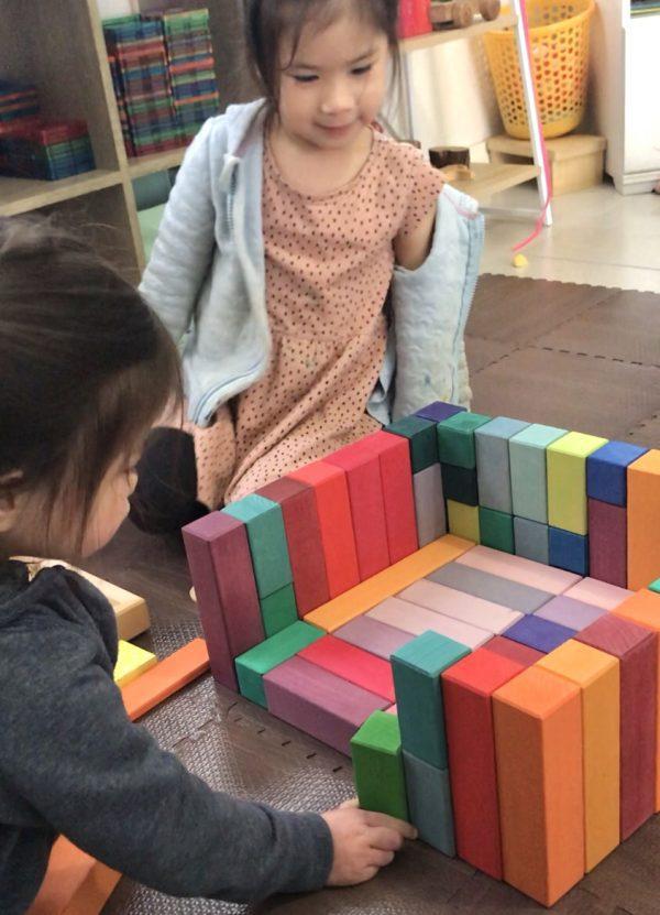 A toddler carefully stacking and lining up wooden blocks to create an enclosed structure, focused on balancing and arranging the pieces during imaginative play. The size of the block allows the toddler to grasp it and develop their gross motor skills