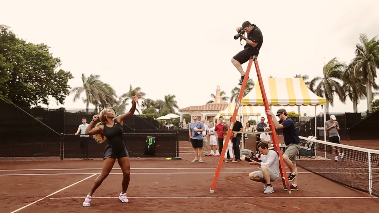 Chase Jarvis high on an A-frame ladder on a clay tennis court, photographing Serena Williams, as a camera-shoot crew and others look on.