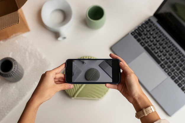 Hands holding a smartphone displaying a ceramic plate on its screen, with a laptop and unpacked items on a table