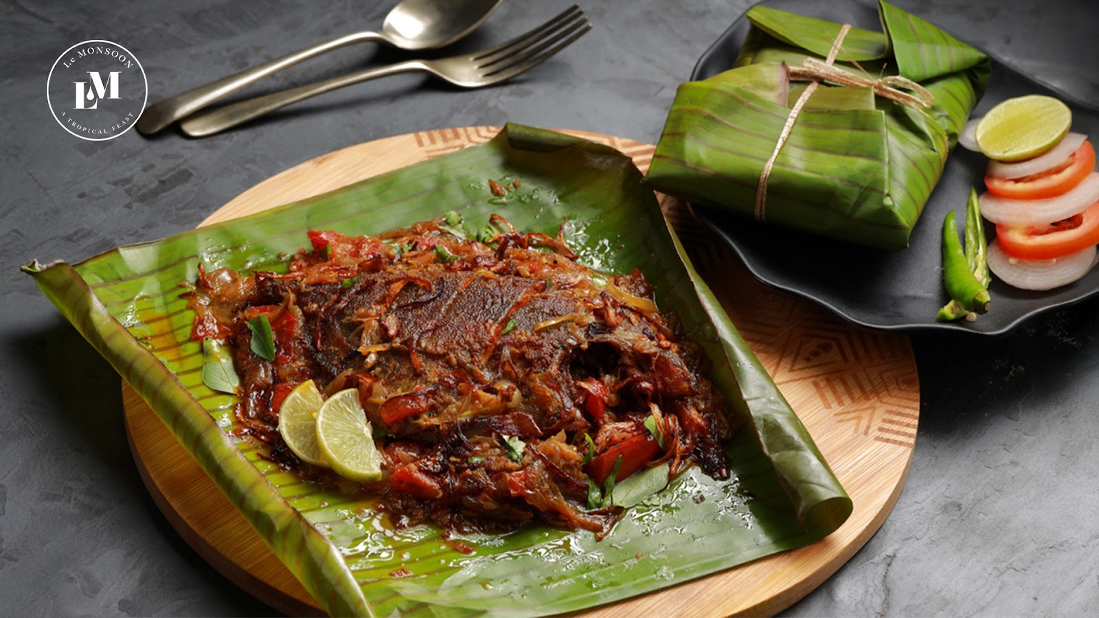A beautifully plated fish dish wrapped in banana leaves, garnished with lime, served on a black plate.