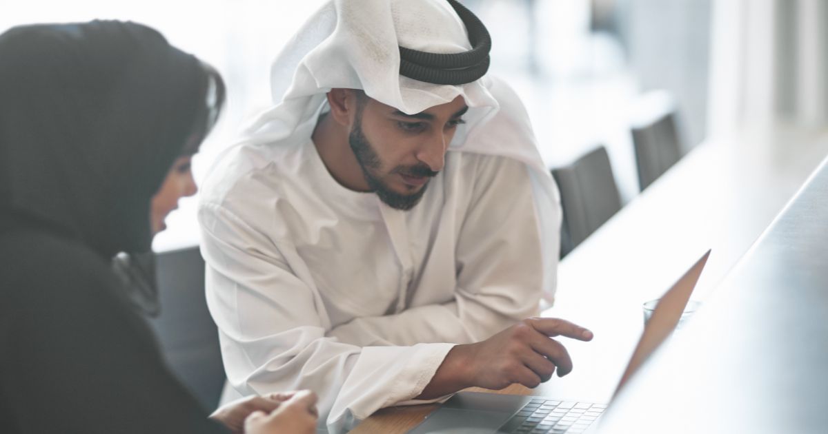 A male Emirati professional wearing a traditional white kandura is engaged in a discussion with a female colleague dressed in a black abaya. They are focused on a laptop screen, with the man pointing towards it, indicating a collaborative work environment. The image illustrates teamwork, business collaboration, and professional consultation, particularly in the context of compliance and workforce development.