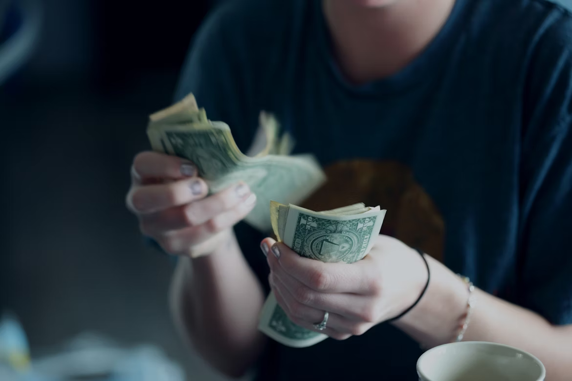 Person wearing a dark shirt counting a stack of one-dollar bills with both hands, partially visible in a softly lit indoor setting.