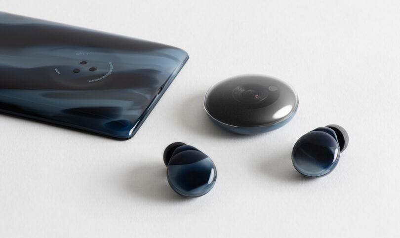 A smartphone, a round camera device, and two wireless earbuds are placed on a white surface. All items have a sleek dark blue design.