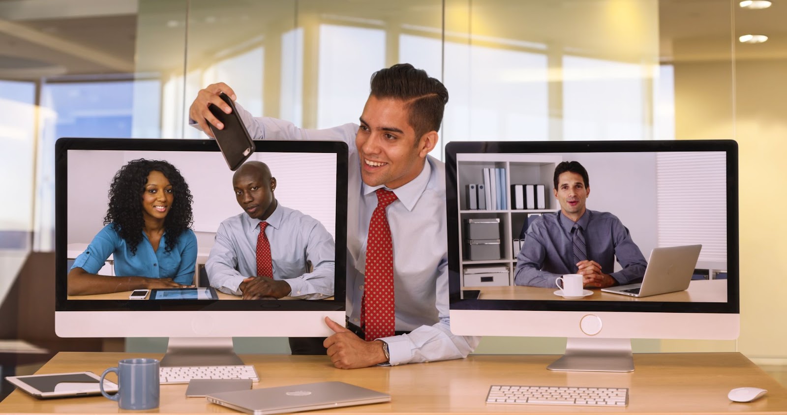 (A man in an office holds a phone toward two computer screens displaying video call participants. One screen shows a woman and man, while the other shows a man sitting at a desk. This Virtual Team Snapshot captures the essence of remote collaboration.)