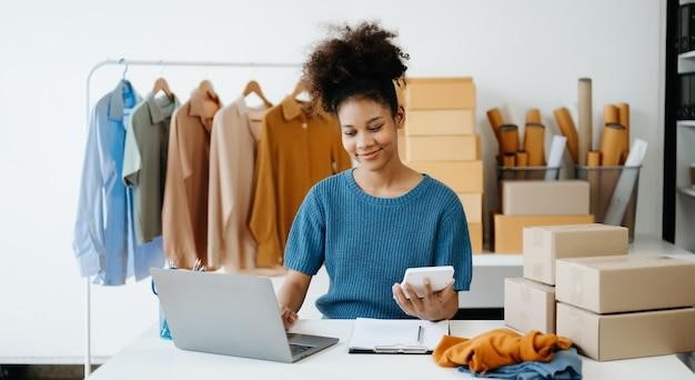 Young African woman running online store Startup small business SME using smartphone or tablet taking receive and checking online purchase shopping order in office