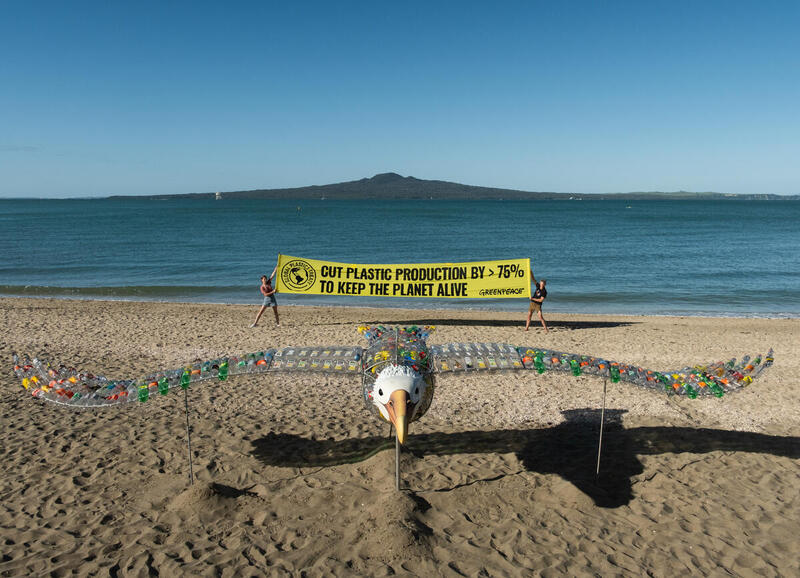 Birdie, the plastic bottle sculpture of a toroa (albatross) is on the beach with Rangitoto in the background. Two people hold a banner saying Cut plastic production by >75% to keep the planet alive. 