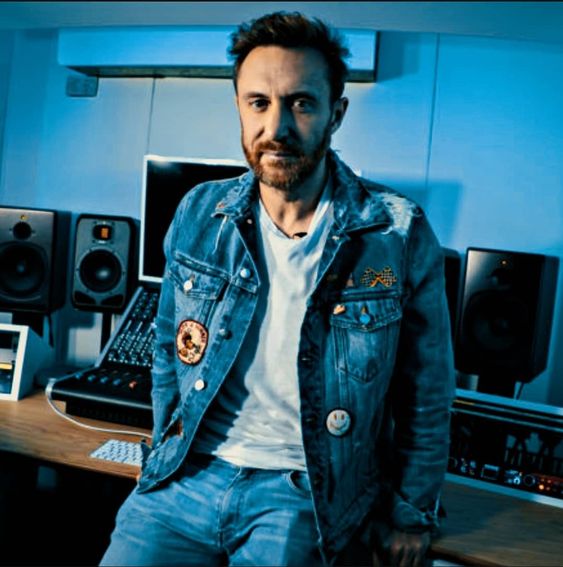 This contains an image of 
David Guetta's sitting in front of a computer desk with speakers on the wall behind him