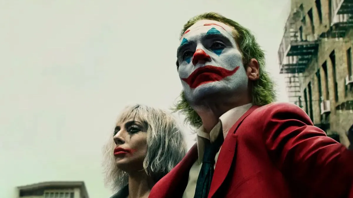 Primer plano del Joker, interpretado por Joaquin Phoenix, con su icónico maquillaje de payaso, acompañado de Harley Quinn, interpretada por Lady Gaga, ambos mirando hacia el horizonte con un fondo de edificios.