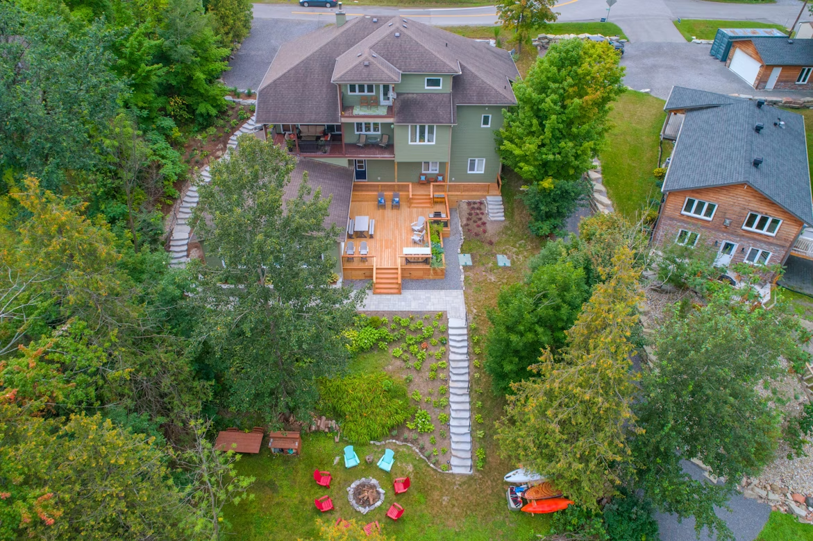 Aerial view of a large backyard with tiered landscaping, wooden deck, stone pathways, lush planting beds, and a circular fire pit area with colorful Adirondack chairs.