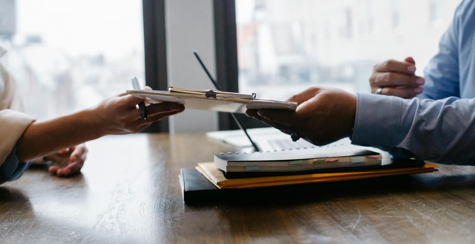 A close-up of two individuals exchanging a clipboard across a table, with notebooks and documents visible in the background, symbolizing the exchange of important information or agreements.