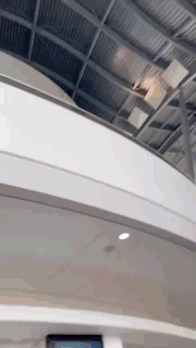 A phone camera smoothly tilts upward inside a spacious, modern event center. The shot captures the high ceiling with exposed metal beams, large windows, and a curved upper level, emphasizing the open and grand architecture of the venue for Atomicon 2025. People are walking into the event and Laura Belgray's introduction slide is shown on a very large monitor mounted from the ceiling of the event space where people are gathering. 