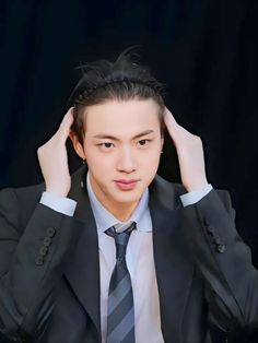 This contains a picture of BTS Jin  in a suit and tie holding his hair up to his head with both hands