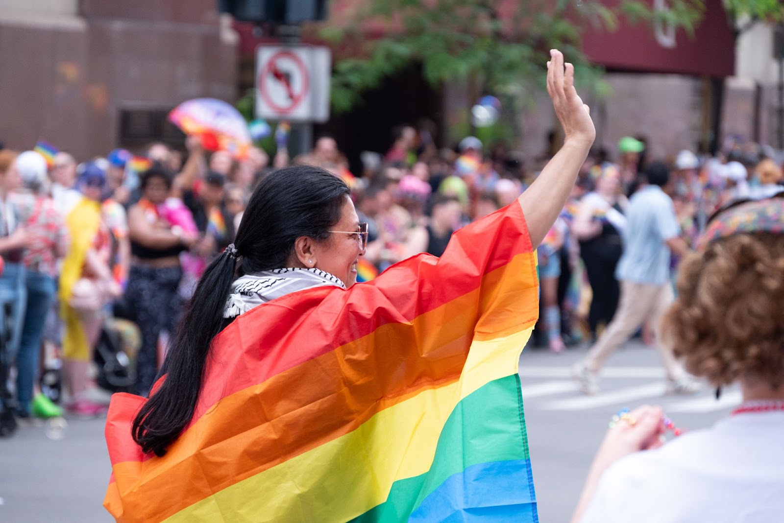 Rep. Tlaib waves while wearing a rainbow flag cape.
