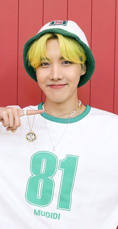 This contain J-Hope with yellow hair wearing a white shirt and green hat is posing for the camera