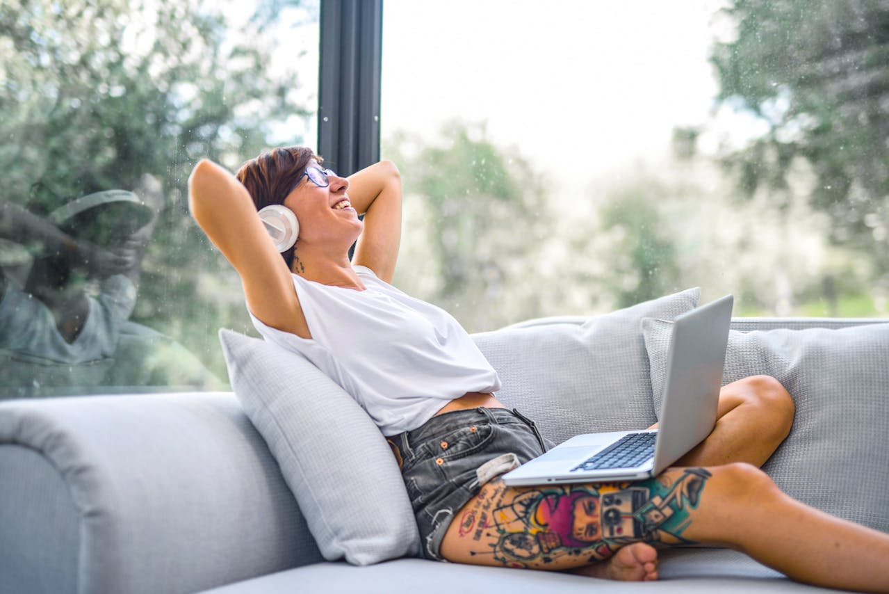 A woman relaxing on a couch with headphones on, enjoying music created using AI, as she has her laptop on her lap in a bright, modern living space.