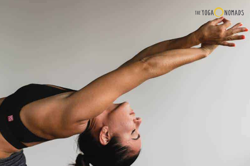 A person is performing a yoga pose on a yoga mat. The individual is in a lunge position with the right leg forward and the left leg extended back, both feet flat on the mat. The person’s arms are raised above the head, with palms facing each other and eyes looking upwards.