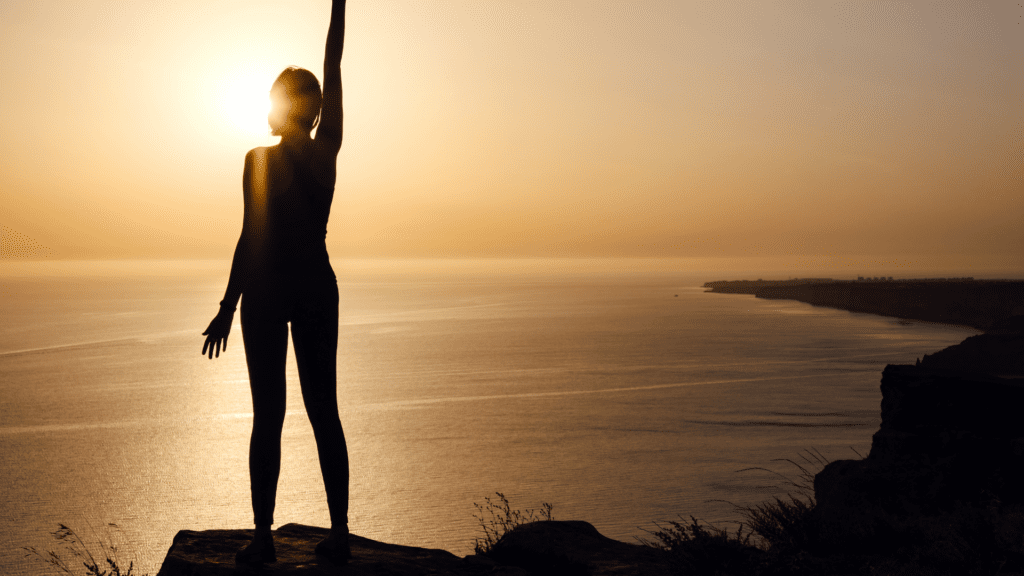 A silhouette of a woman standing on a cliff at sunset, raising one arm towards the sky, representing empowerment and personal freedom.