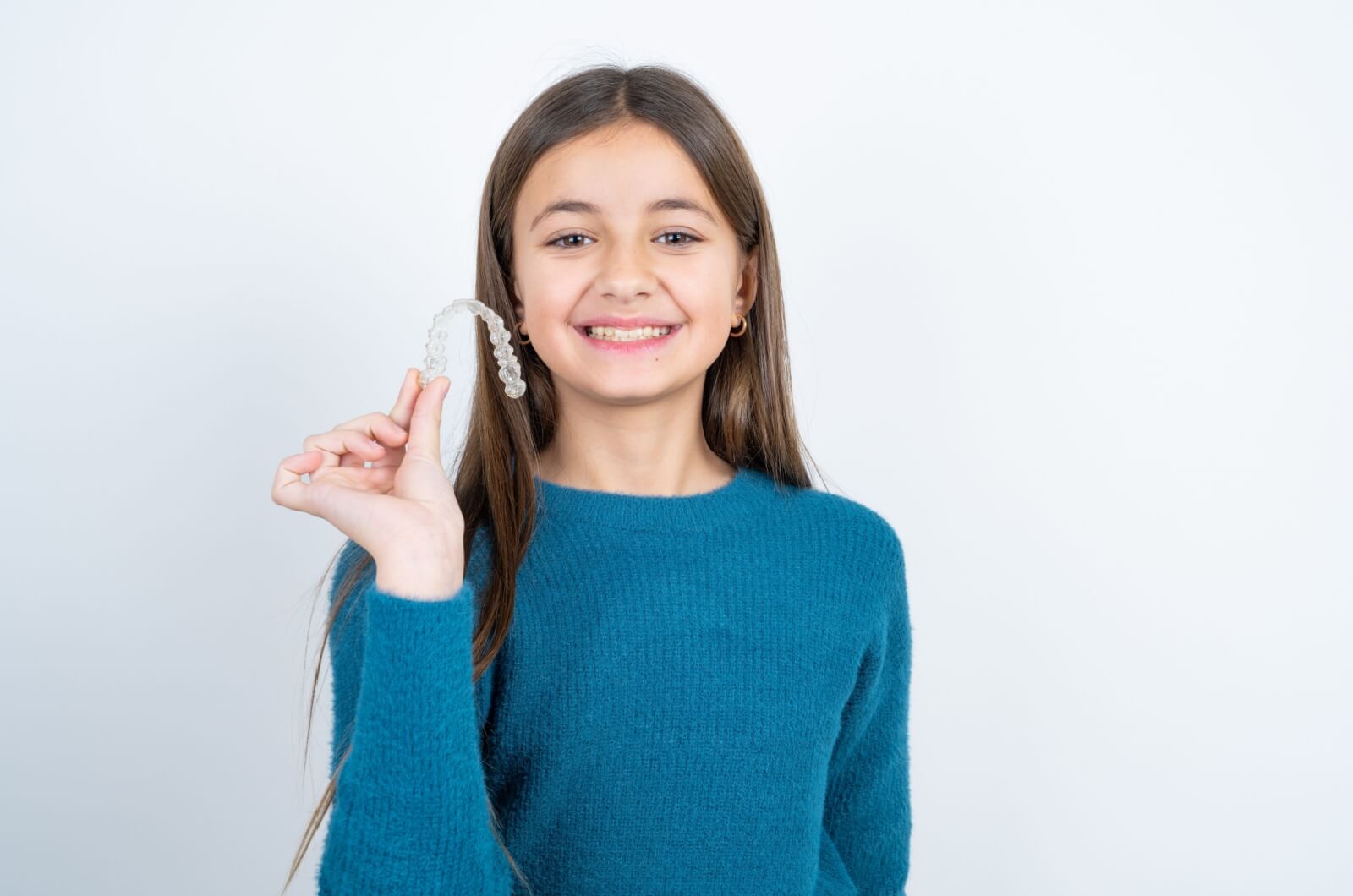 A smiling young girl in a blue sweater holding up a clear aligner in her right hand.