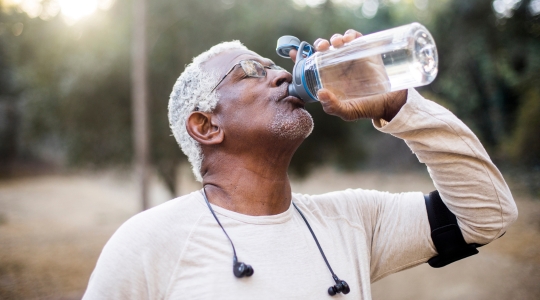 An elderly man with gray hair drinks water from a large plastic bottle while on an outdoor walk. He is wearing a long-sleeved shirt and has earphones around his neck.