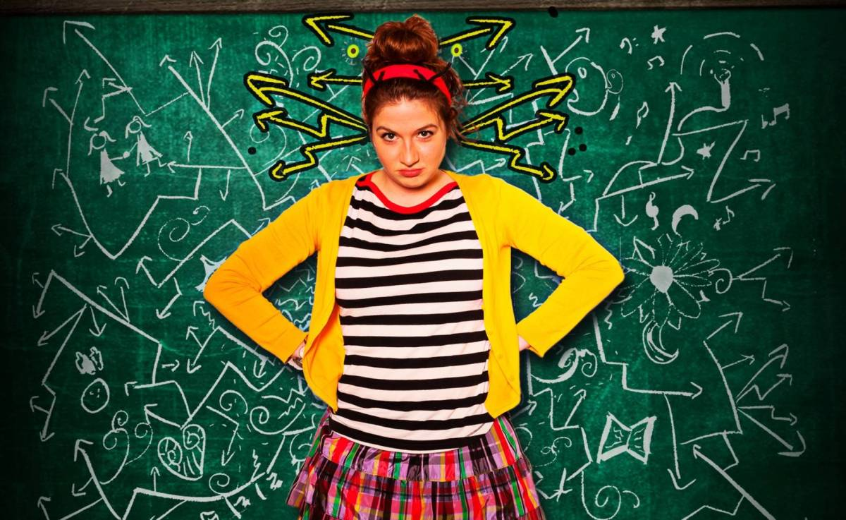 A white woman stands in front of a green blackboard covered in squiggles