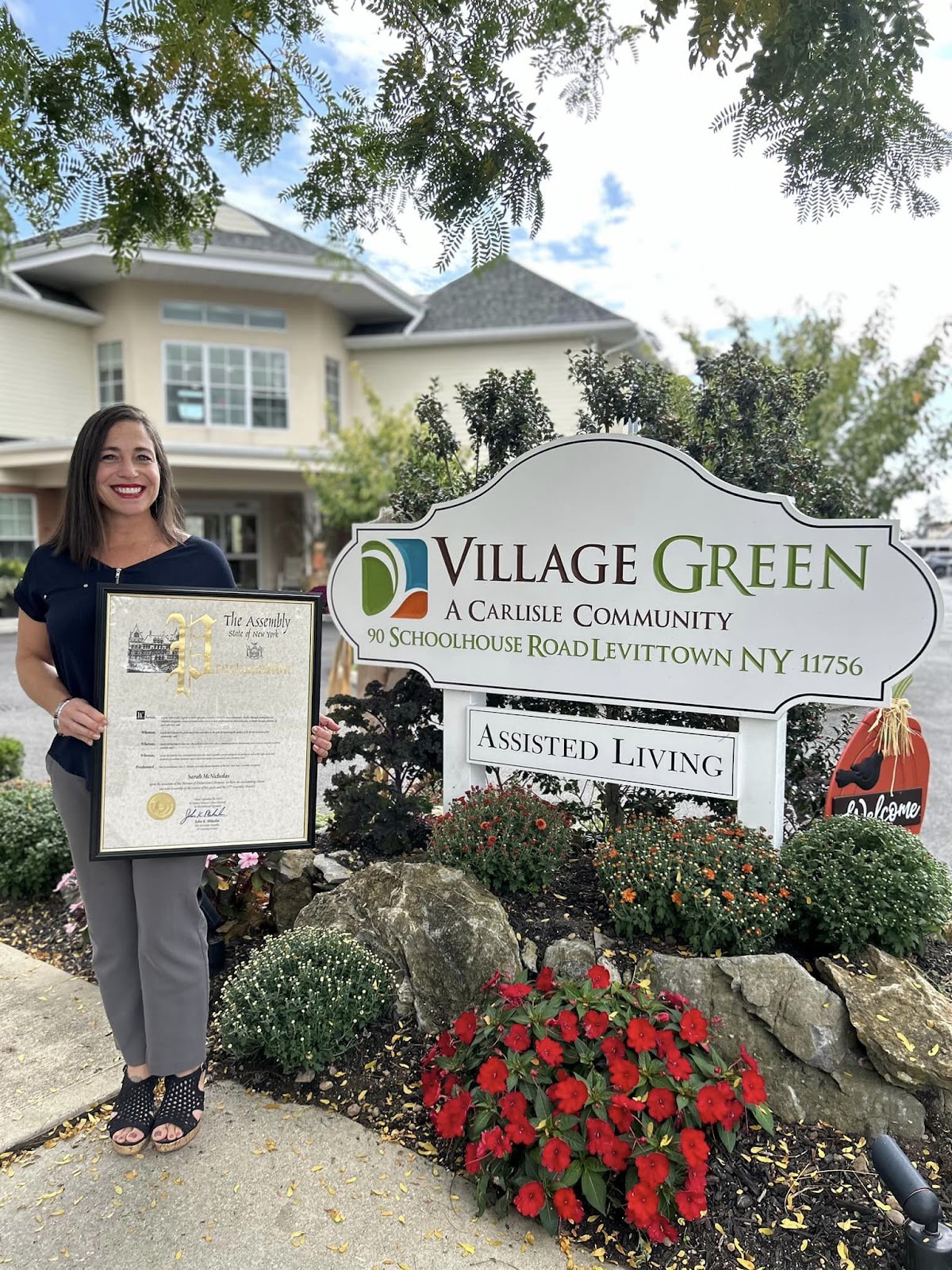 A smiling staff member with a framed certificate in her hands in front of an outdoor Village Green sign