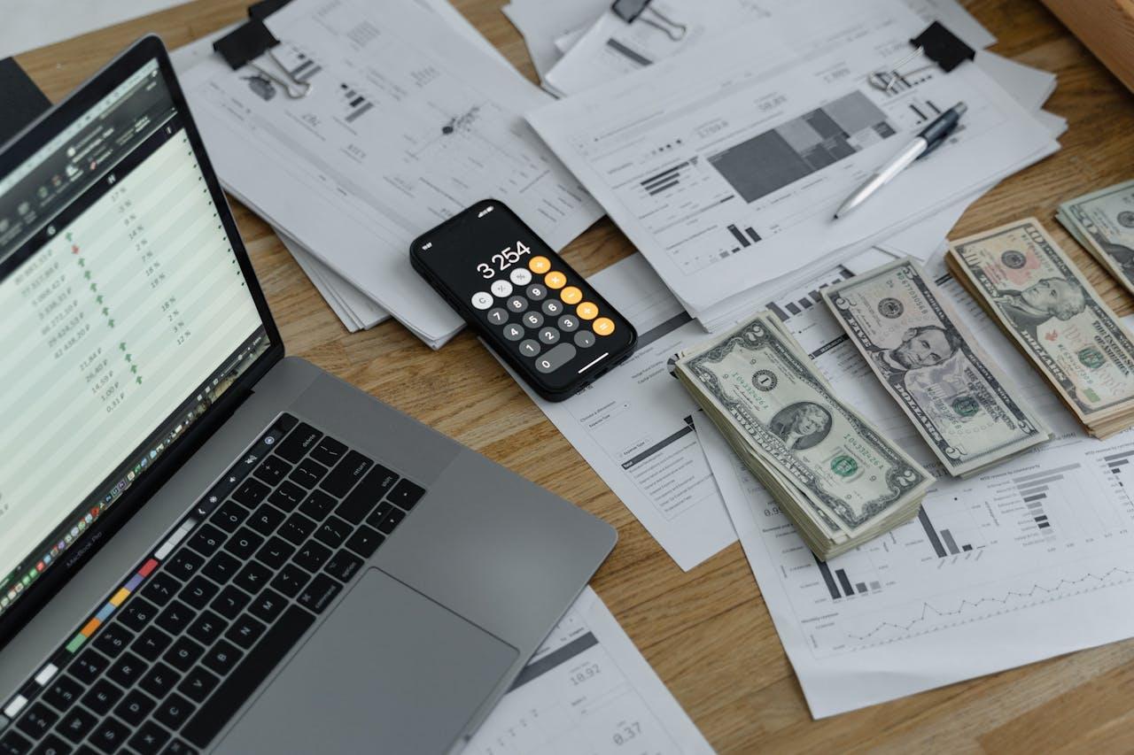 A laptop, calculator, papers, and dollar bills on a wooden desk