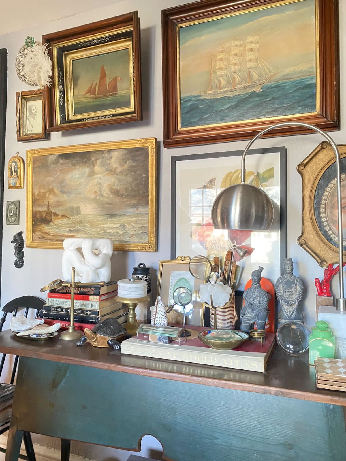 A collection of framed nautical-themed paintings (depicting ships at sea) hangs on the wall above a wooden table. The table is adorned with various decorative items, including books, statues, a lamp, and small artifacts.