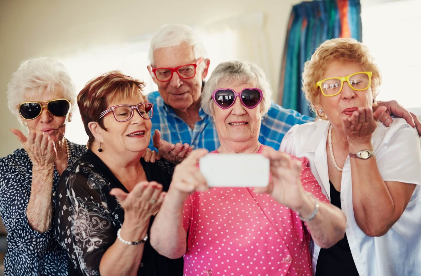 A group of seniors wearing funny eyeglasses and sunglasses gather around a phone to take a selfie