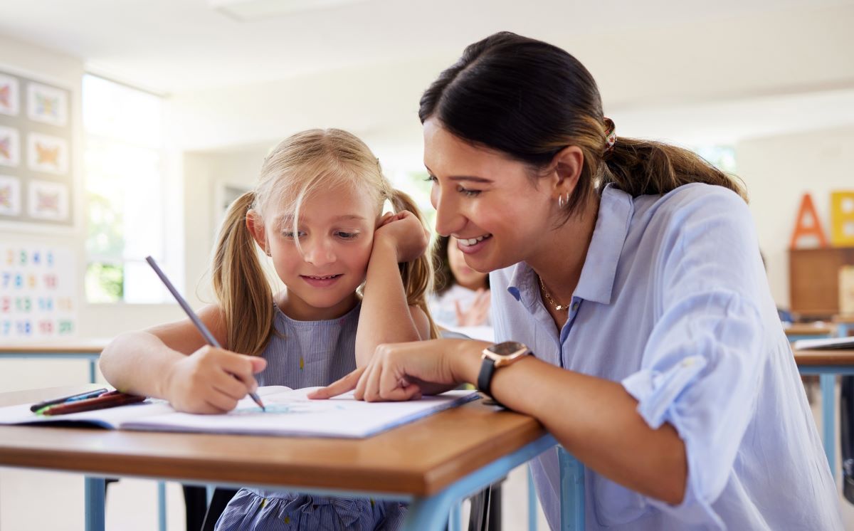 Etapele cheie ale dezvoltarii emotionale la copii educatoare ajuta zambind o fetita la rezolvarea exercitiilor - babymatters.ro