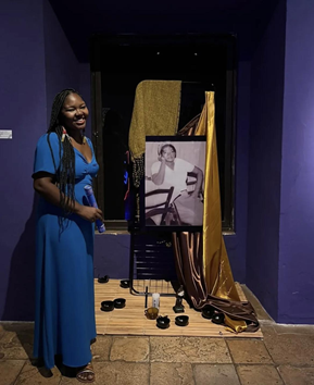 Uma mulher negra, de tranças longas e vestido azul sorri. Ela está ao lado do retrato de sua avó. Roberta é uma Agente Cultura, participando do mapeamento cultural do projeto Brasil Memória em Rede II do Museu da Pessoa.
