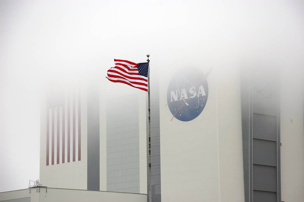 the top of a large building with the American flag on one side and the NASA insignia on another side is covered by fog