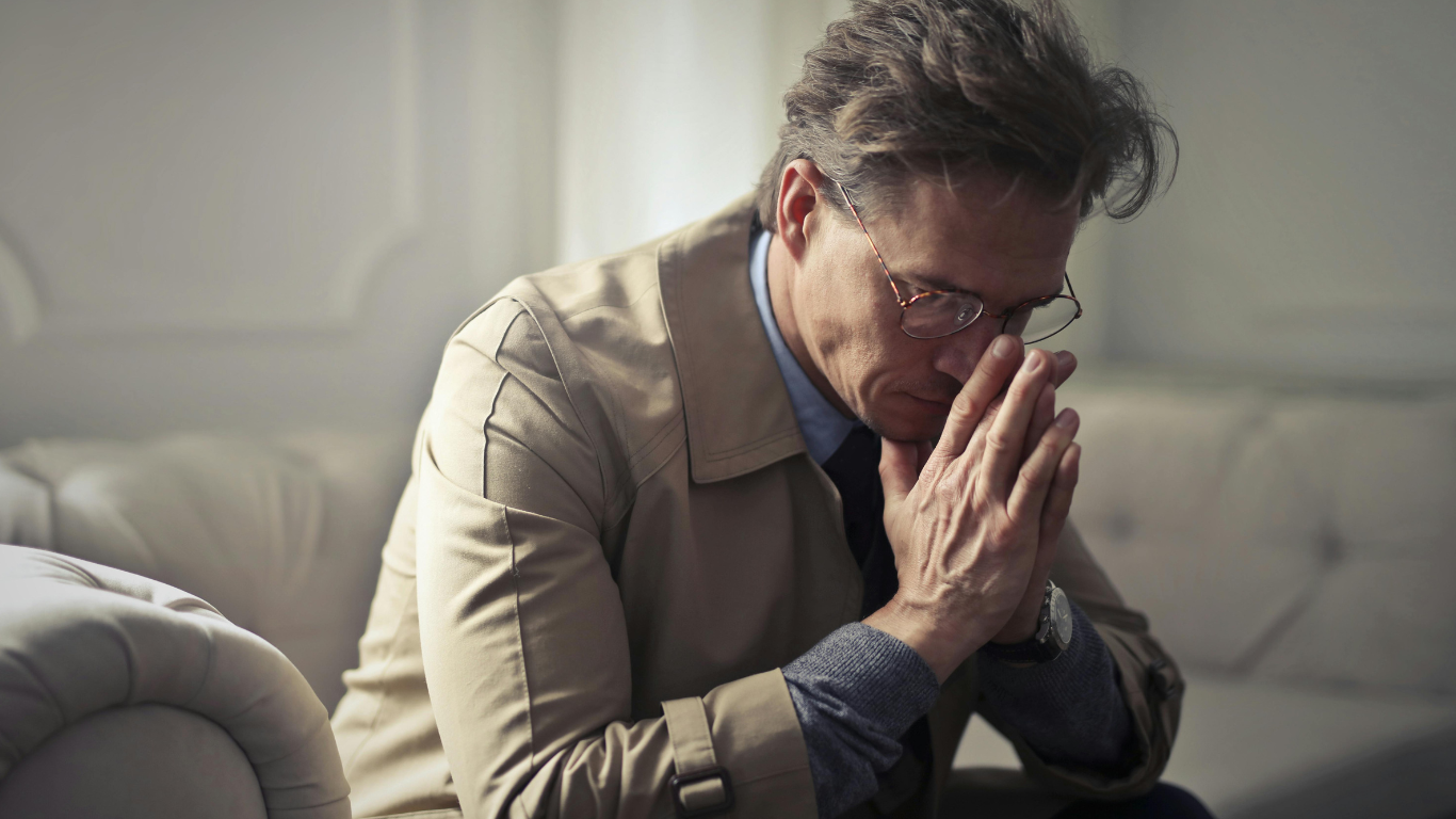 Middle-aged man sitting on a couch, appearing deep in thought or emotionally distressed, with hands clasped in front of his face.