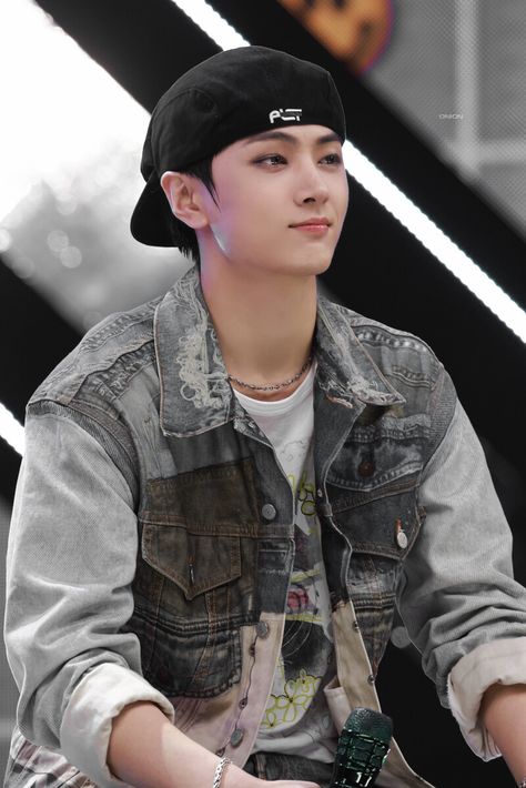 This contain an image of  ENHYPEN‘s Jay wearing a denim jacket and cap sitting on a chair with his hands in his pockets