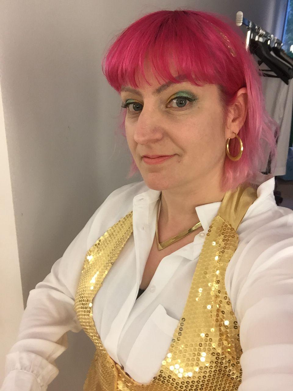 Picture of a woman with pink hair, wearing a gold sequin vest, a white shirt. She is looking very happy.