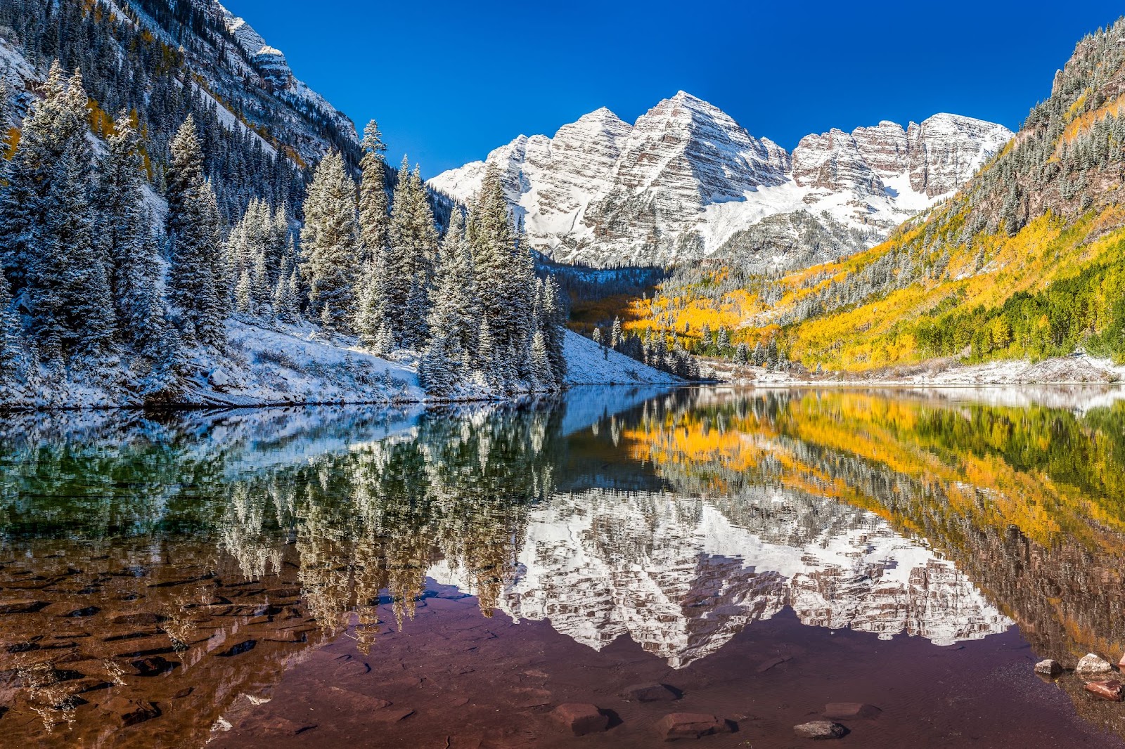 Crystal-clear Maroon Lake in Aspen, Colorado, reflecting the snow-covered Maroon Bells, evergreen trees and vibrant yellow autumn foliage under a bright blue sky.