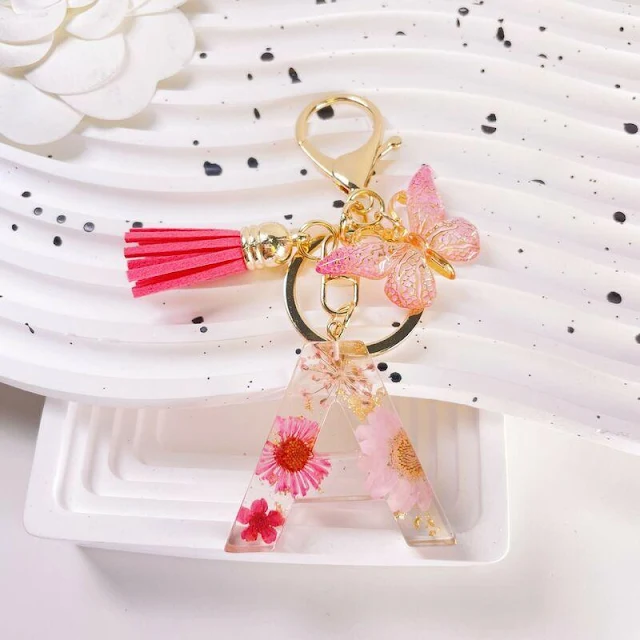 Handmade Floral Initial Keychain with Butterfly & Tassel A clear epoxy resin keychain in the shape of the letter 'A', embedded with delicate pink dried flowers and flecks of gold. It's attached to a gold-tone keyring along with a pink tassel charm and two light pink butterfly charms. The keychain is displayed against a white textured background with black speckles.