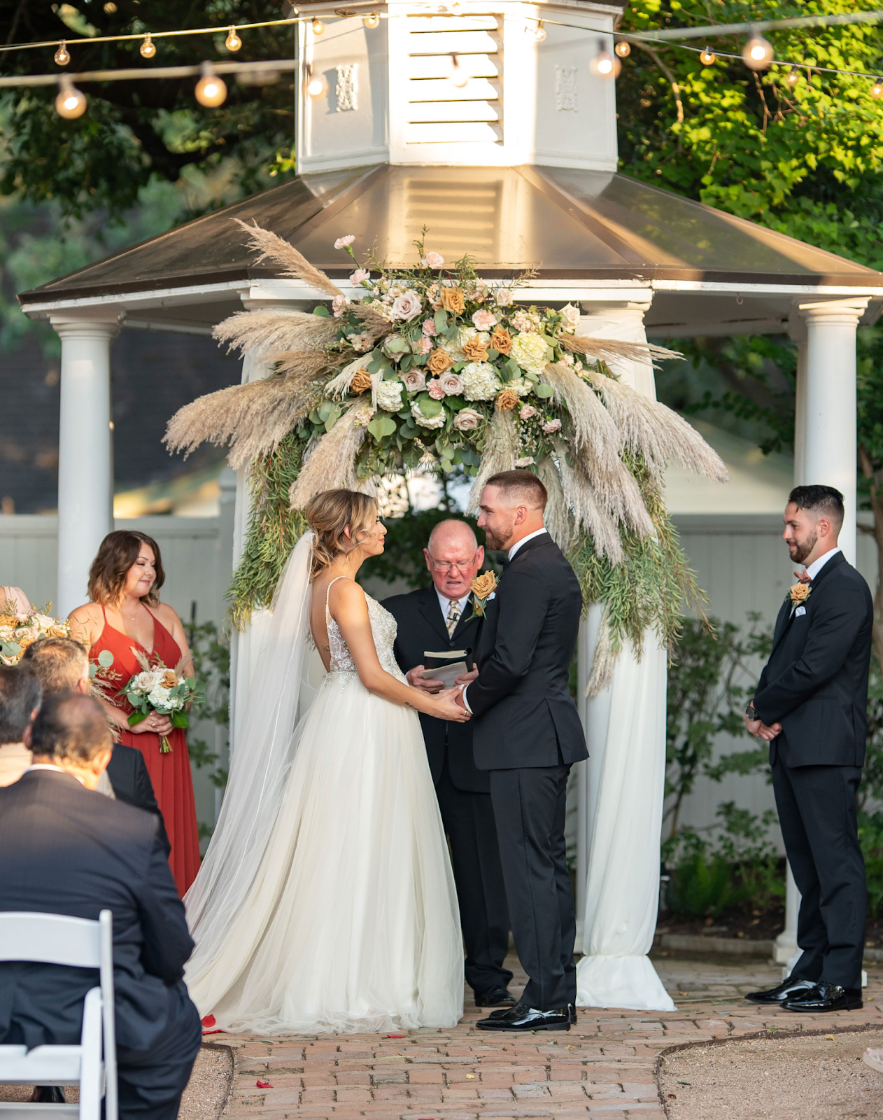 Bride and groom exchange vows in front of a gazebo adorned in Boho Chic style during a garden wedding. The gazebo features a large floral arrangement with pampas grass and greenery, complemented by soft drapery elegantly draped around its columns, creating a romantic and stylish atmosphere at Butler’s Courtyard in Galveston County Texas.