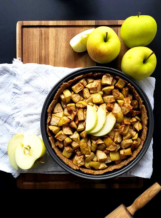 Tarte aux pommes garnie de morceaux de pommes cuites, avec des tranches de pomme et des pommes fraîches sur une planche en bois.